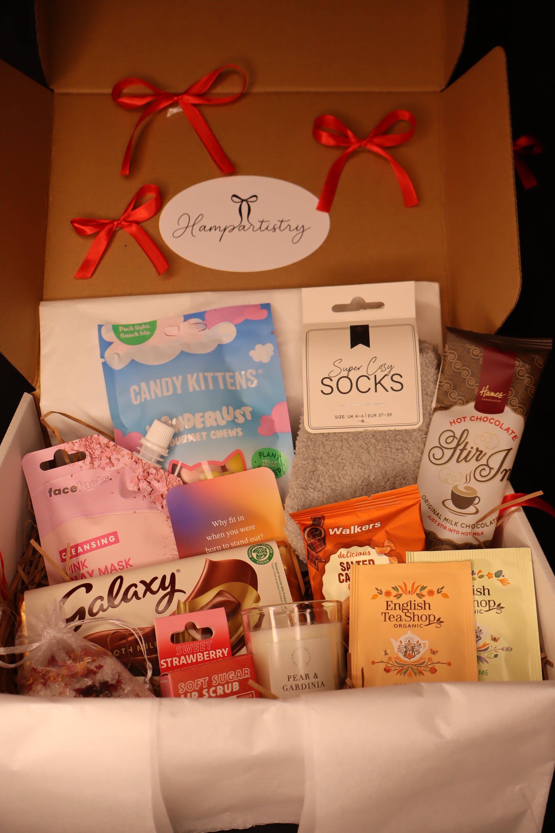 Gift box with various items including socks, candy, and a bottle, with a red ribbon and label.