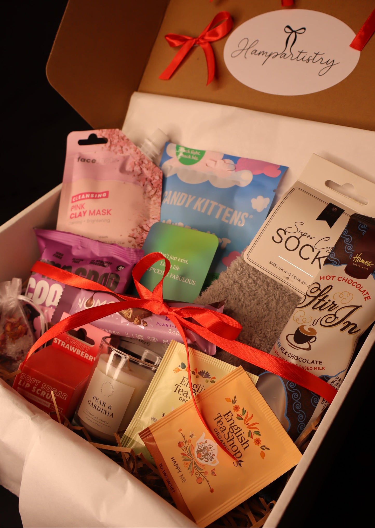 Gift box with various items including candy kittens, a scented candle, and a face mask, tied with a red ribbon.