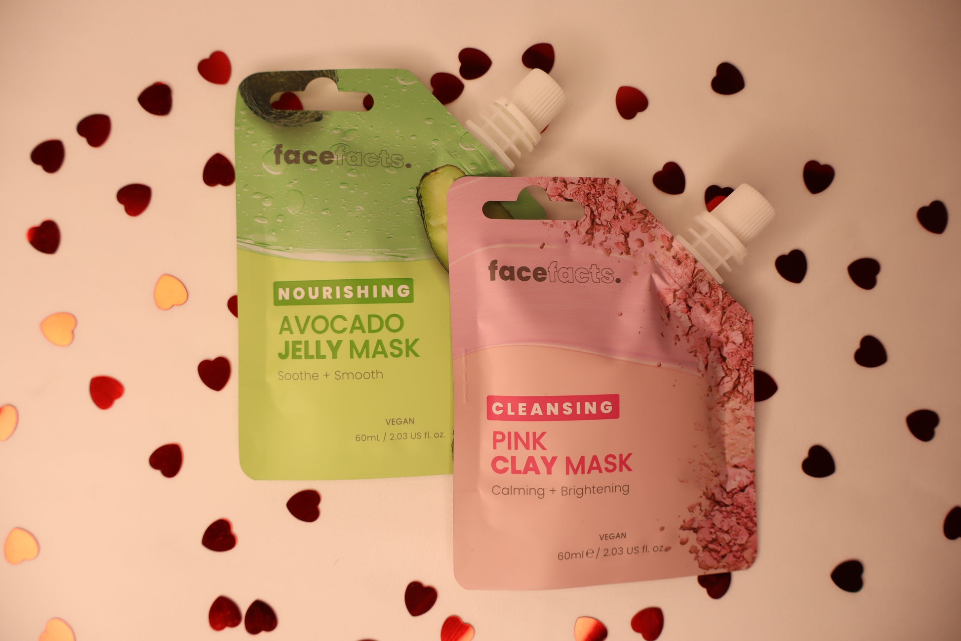 Two face mask packages, one green and one pink, on a white background with heart confetti.