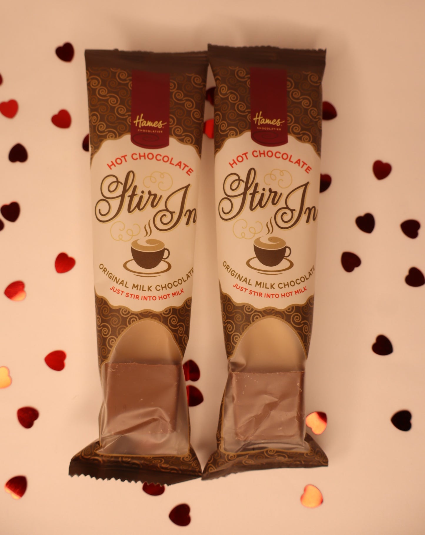 Two packages of Hames Stir In Hot Chocolate on a decorative background with heart shapes.