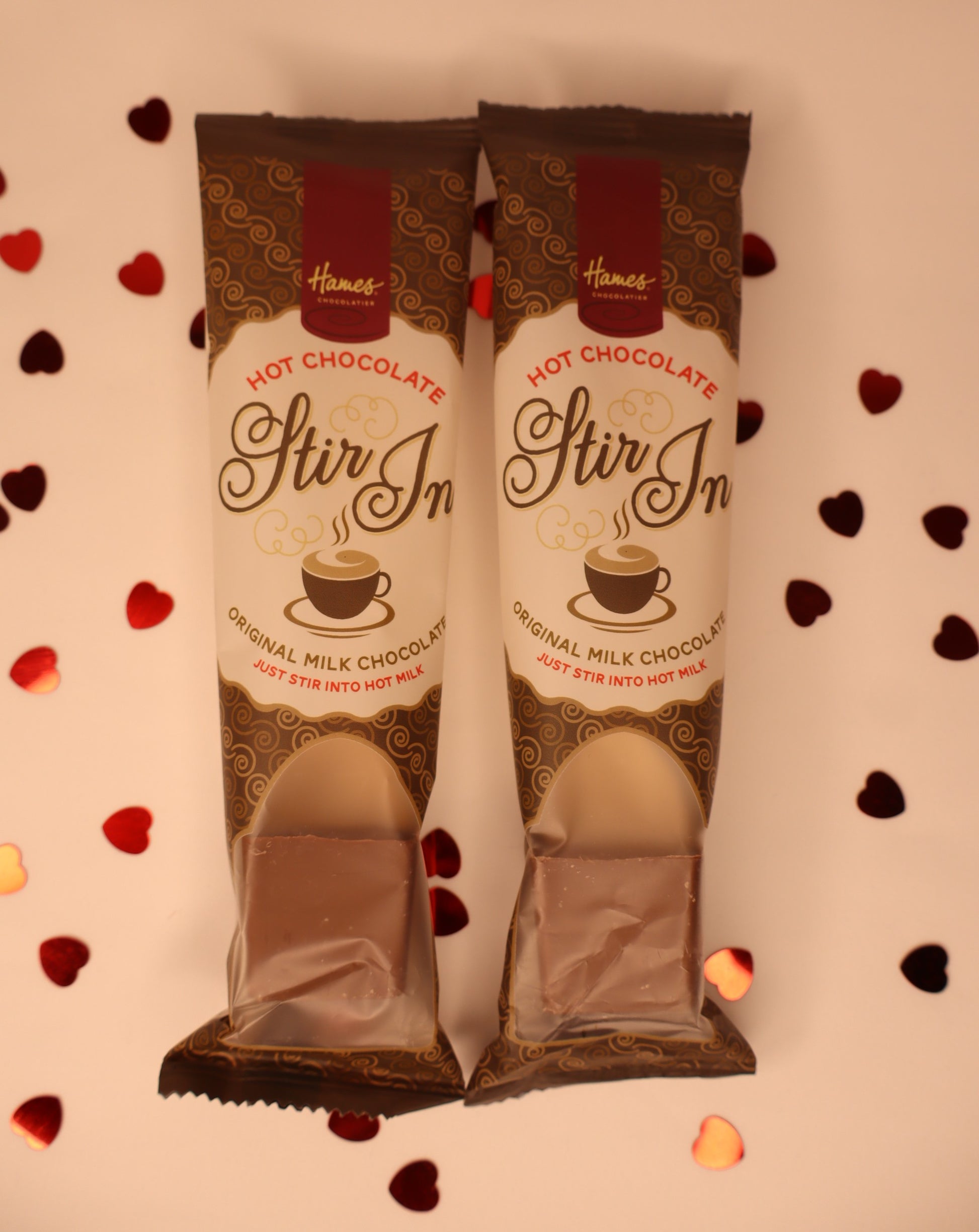 Two packages of Hames Stir In Hot Chocolate on a decorative background with heart shapes.