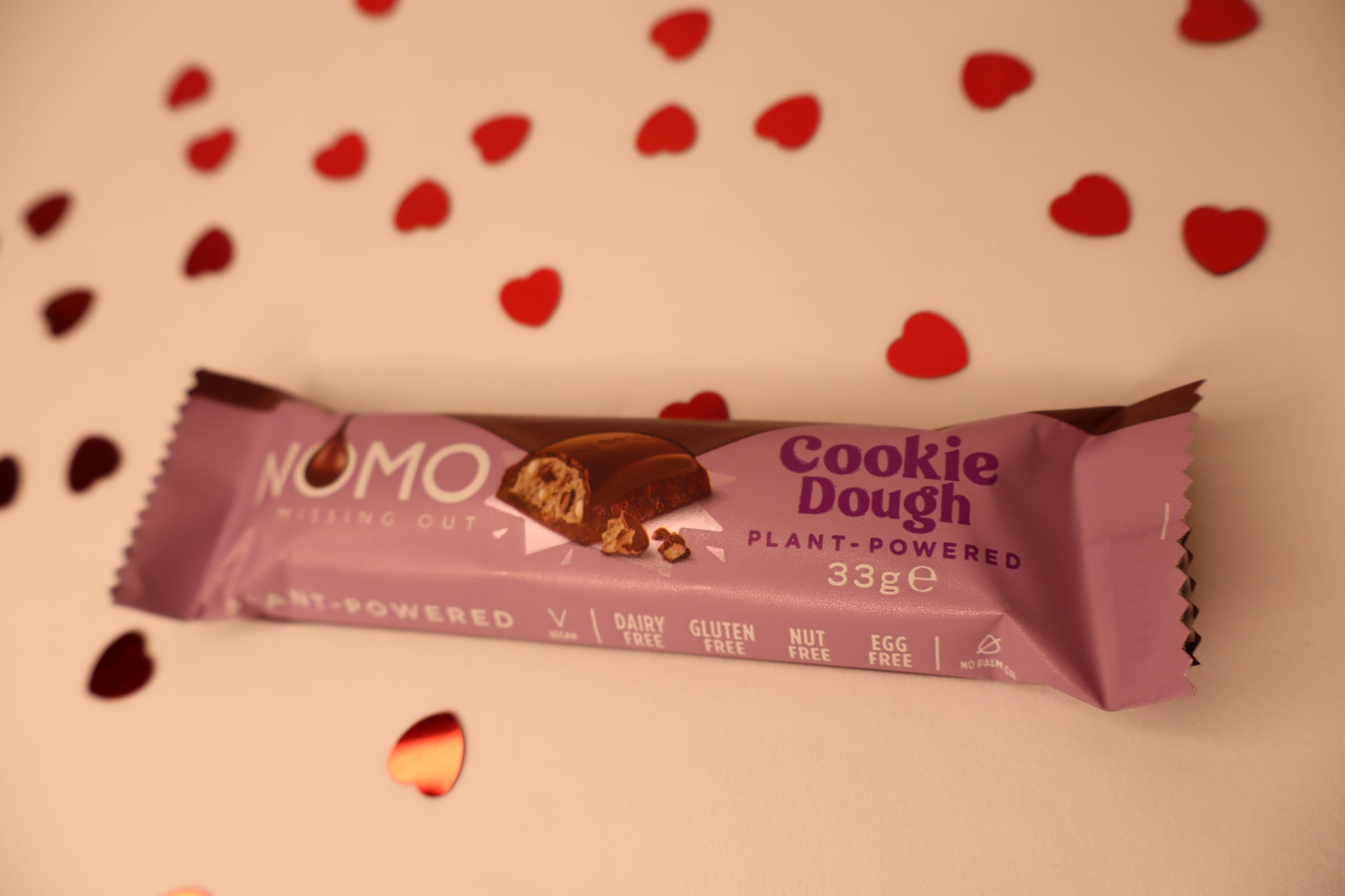 NOMO Cookie Dough bar with purple packaging on a white background with heart shapes.